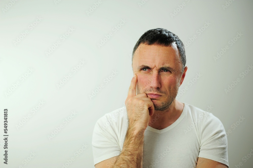 Problems of experiencing a decision Strict unshaven man looks into the frame Hold your chin with your hand and think He has a strong piercing blue eyes He is wearing a white T-shirt white background Problems of experiencing a decision Strict unshaven man looks into the frame Hold your chin with your hand and think He has a strong piercing blue eyes He is wearing a white T-shirt white background