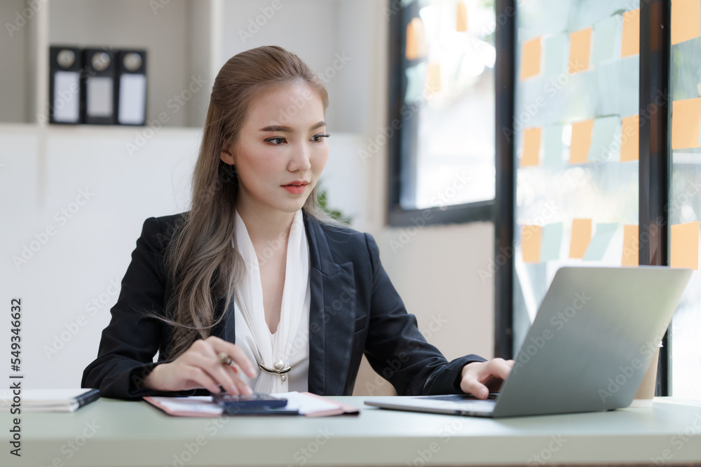 Young attractive Asian business woman with messy desk in office, sitting at the desk, working at contemporary office and documents. Using calculator. Young attractive Asian business woman with messy desk in office, sitting at the desk, working at contemporary office and documents. Using calculator.