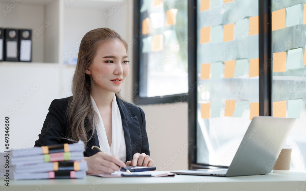 Young attractive Asian business woman with messy desk in office, sitting at the desk, working at contemporary office and documents. Using calculator. Young attractive Asian business woman with messy desk in office, sitting at the desk, working at contemporary office and documents. Using calculator.