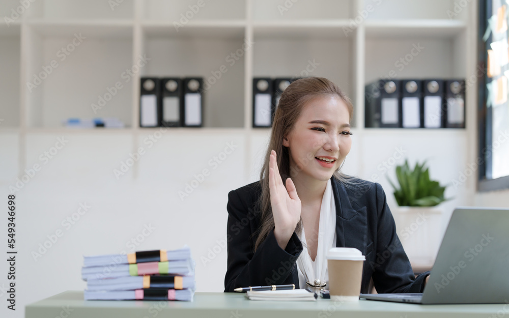 Young attractive Asian business woman with messy desk in office, sitting at the desk, working at contemporary office and documents. hand wave, video call on laptop Young attractive Asian business woman with messy desk in office, sitting at the desk, working at contemporary office and documents. hand wave, video call on laptop