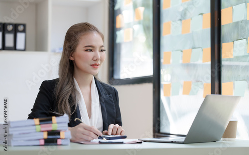 Young attractive Asian business woman with messy desk in office, sitting at the desk, working at contemporary office and documents. Using calculator.