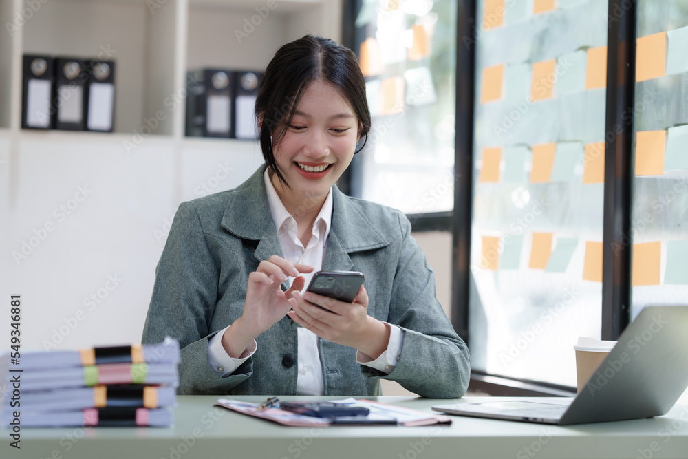 Pretty Asian business woman in black suit working at her office desk, with document file and paperwork. texting mobile smartphone or chat message Pretty Asian business woman in black suit working at her office desk, with document file and paperwork. texting mobile smartphone or chat message