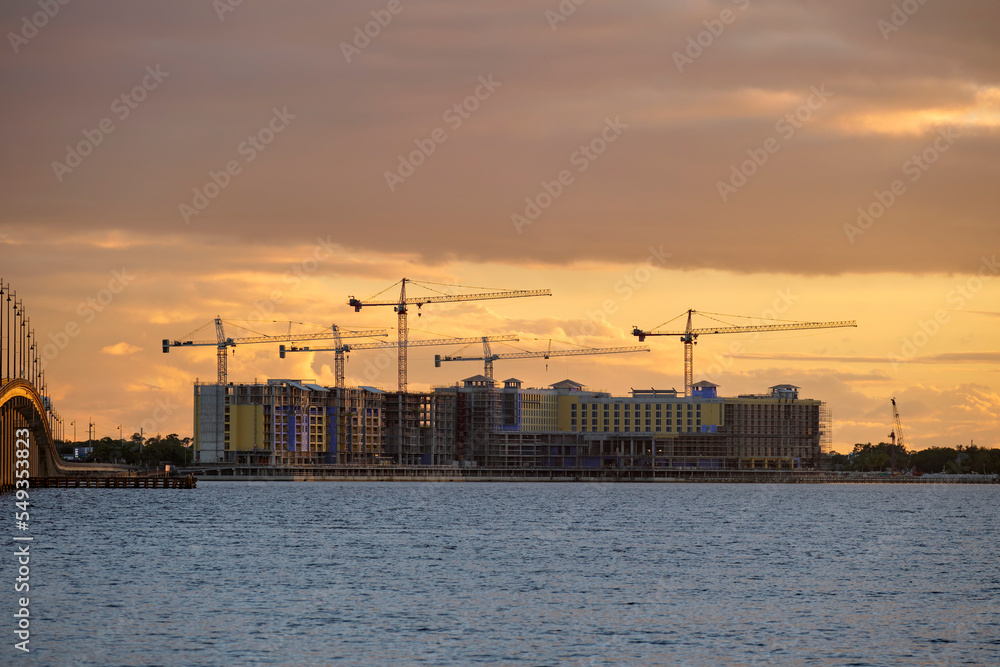 Obraz premium Tower cranes and frame structure of high residential apartment buildings at construction site on sea bay shore. Real estate development at waterfront