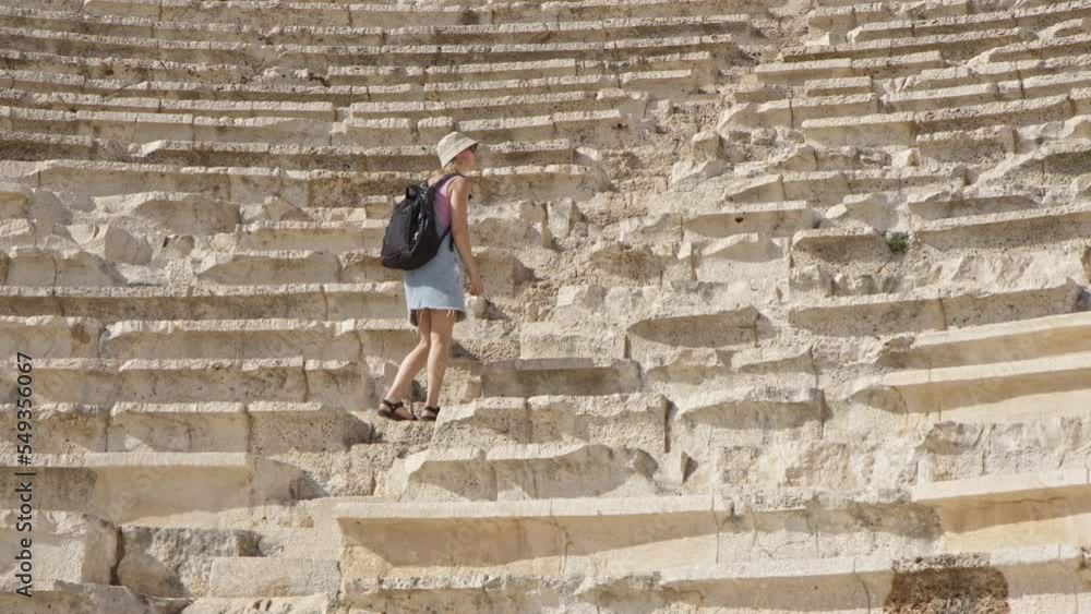 young woman in panama hat and denim overalls climbs stone stairs of antique amphitheater. traveler girl in her journey climbs up historical monument in ancient city in open air. young woman in panama hat and denim overalls climbs stone stairs of antique amphitheater. traveler girl in her journey climbs up historical monument in ancient city in open air.