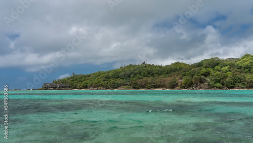 The island in the turquoise ocean is completely overgrown with tropical vegetation. The villas of the hotel are visible among the trees. Blue sky with clouds. Seychelles
