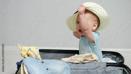 Excited baby girl sits in suitcase with clothes and gets ready for summer vacation. Child takes off straw hat and looks around. Kid wants to help to prepare