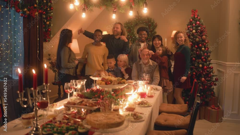 Asian woman sets timer on camera or phone. Happy multi cultural family takes group photo. They celebrating Christmas or New Year. Table with dishes and candles. Family Christmas dinner. Camera view. Asian woman sets timer on camera or phone. Happy multi cultural family takes group photo. They celebrating Christmas or New Year. Table with dishes and candles. Family Christmas dinner. Camera view.