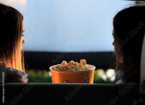 Two female friends with popcorn sitting in the car while watching a movie at drive in cinema. Selective focus. Entertainment, leisure activities, hobby concept