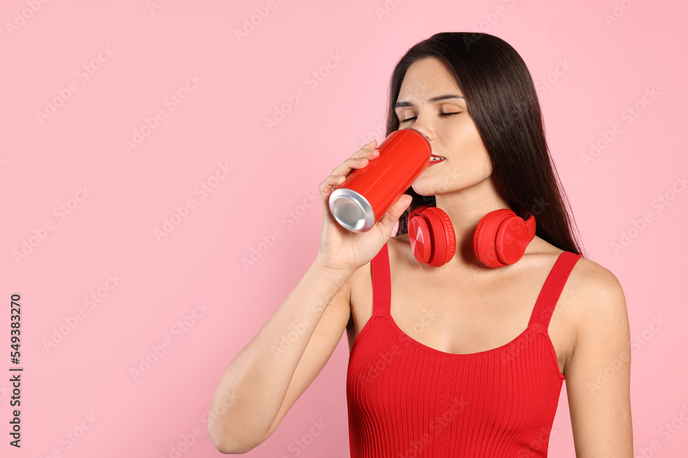 Obraz premium Beautiful young woman with headphones drinking from tin can on pink background. Space for text