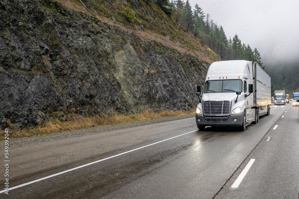 Big rig powerful white semi truck transporting cargo in refrigerator semi trailer running on the wet slippery road with fog. Big rig powerful white semi truck transporting cargo in refrigerator semi trailer running on the wet slippery road with fog.