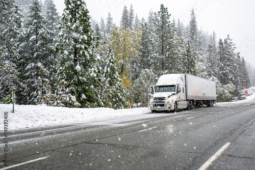 Big rig white semi truck with loaded dry van semi trailer standing on the road shoulder of a winter highway during a snow storm near Shasta Lake in California waiting for favorable weather