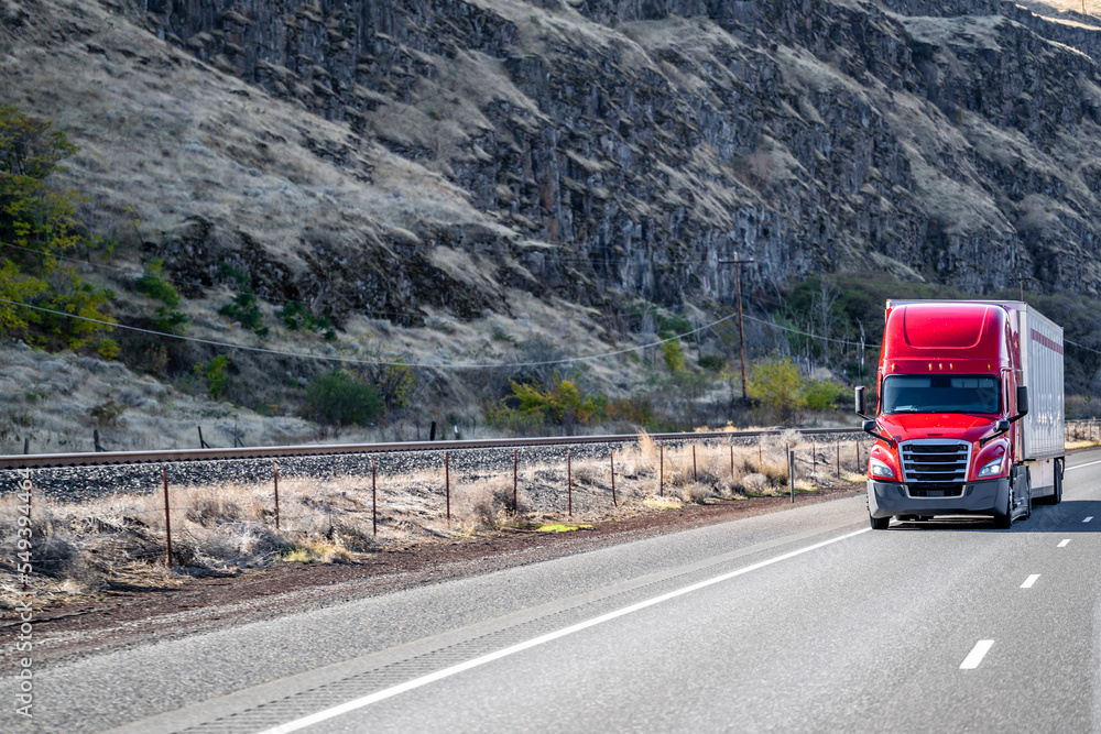 Bright red big rig long hauler semi truck with dry van semi trailer running on the road along the rocky mountain range