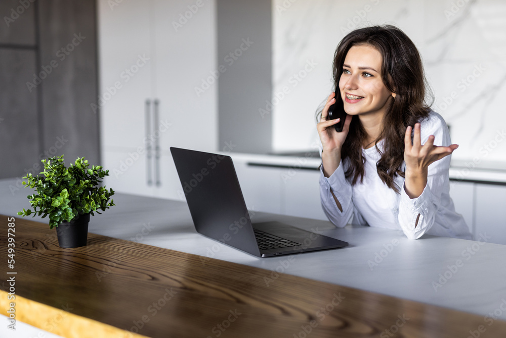 Photo of a young cheerful woman indoors at home at the kitchen using laptop computer talking by mobile phone. Photo of a young cheerful woman indoors at home at the kitchen using laptop computer talking by mobile phone.