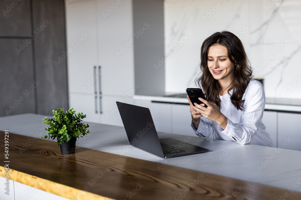 Young woman having breakfast while sitting at the kitchen table, working on laptop computer, using mobile phone Young woman having breakfast while sitting at the kitchen table, working on laptop computer, using mobile phone