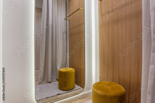 Fitting room in a modern store with a large mirror and a small brown pouf.