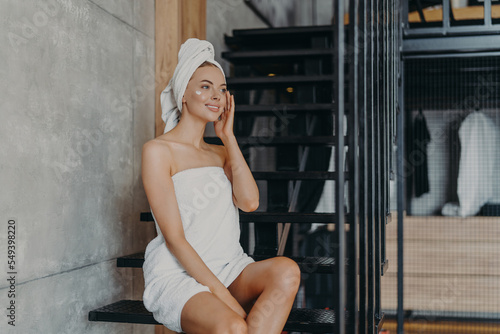 Quadro em tela Pensive beautiful young woman applies body cream, enjoys softness of skin, sits on stairs at home, wrapped in white bath towel, looks thoughtfully into distance