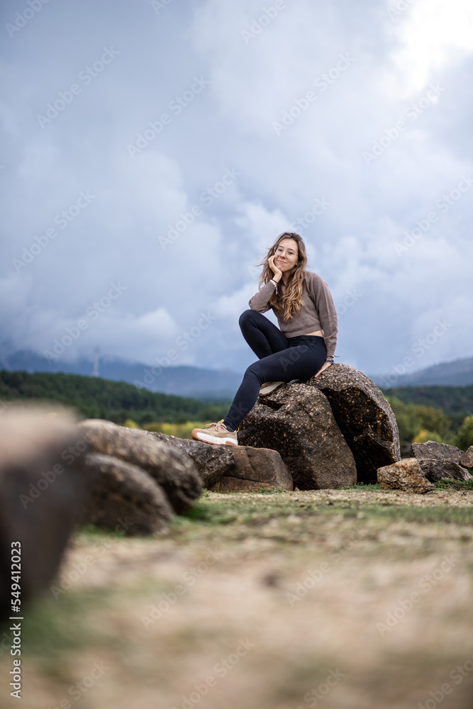young woman sitting on rocks with a beautiful green landscape with water and smiling at the camera, young beautiful girl sitting on a rock in the middle of a lake with a mountain landscape background 
