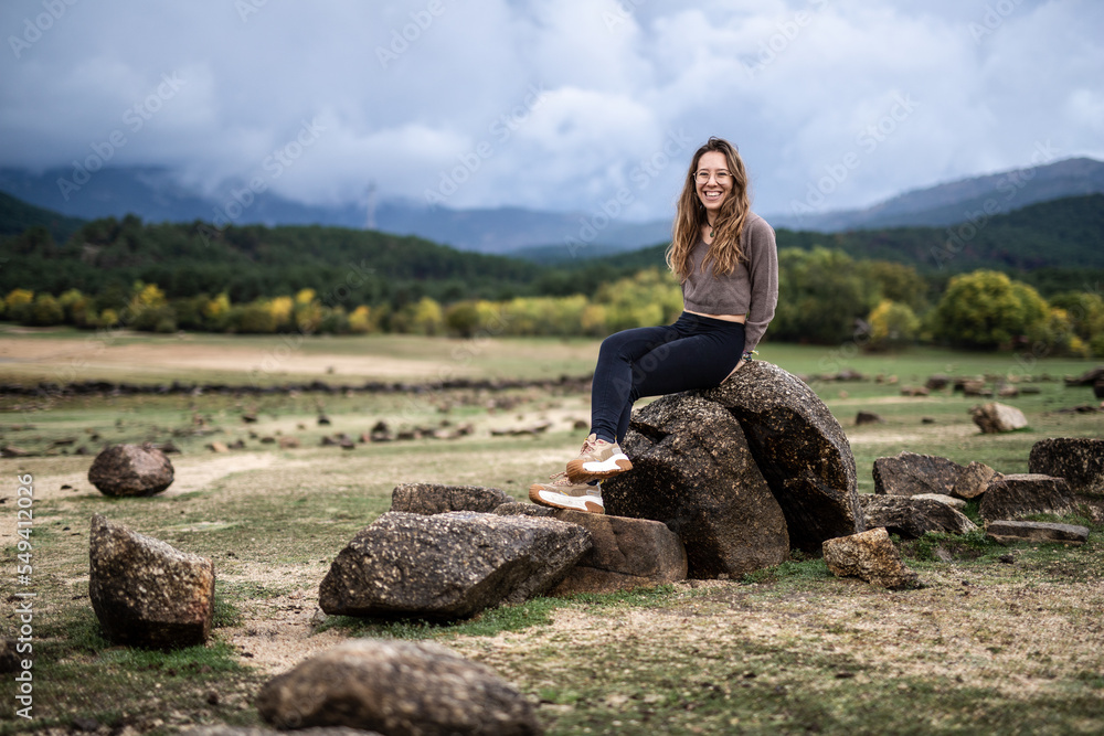 Young beautiful girl sitting on a rock in the middle of a lake with a mountain landscape background smilling at the camera in winter season Young beautiful girl sitting on a rock in the middle of a lake with a mountain landscape background smilling at the camera in winter season