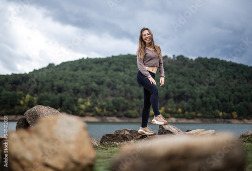 Young beautiful girl on a rock in the middle of a lake with a mountain landscape background smilling at the camera in winter season