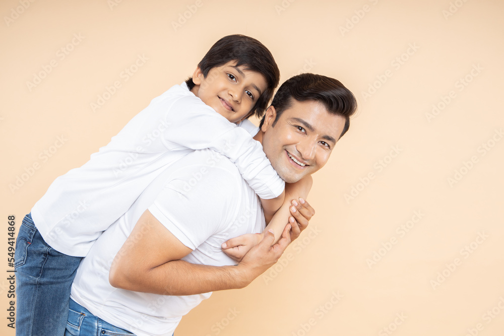 Happy young indian father and son wearing white casual t-shirt standing over isolated beige background. Fatherhood and family bonding Happy young indian father and son wearing white casual t-shirt standing over isolated beige background. Fatherhood and family bonding
