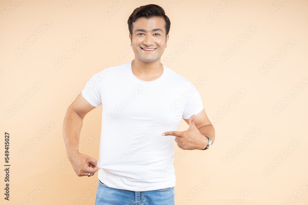 Handsome young indian man wearing white casual t-shirt pointing finger at blank space. front view of a mockup design print. Handsome young indian man wearing white casual t-shirt pointing finger at blank space. front view of a mockup design print.