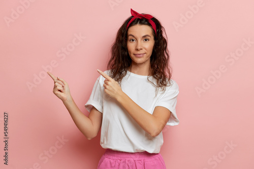 Pretty and positive young woman shows fingers a side to copy space, stands isolated over pink background