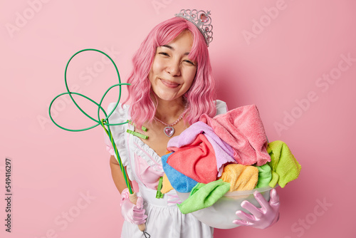 Horizontal shot of pleased Asian woman with pink hair does housework poses with laundry and carpet beater glad almost finish all her tasks dressed like housemaid isolated over rosy background photo