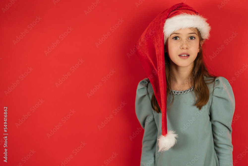 Portrait of a girl with a surprised look in the cap of a santa on a red background, the concept of Christmas, copy space Portrait of a girl with a surprised look in the cap of a santa on a red background, the concept of Christmas, copy space