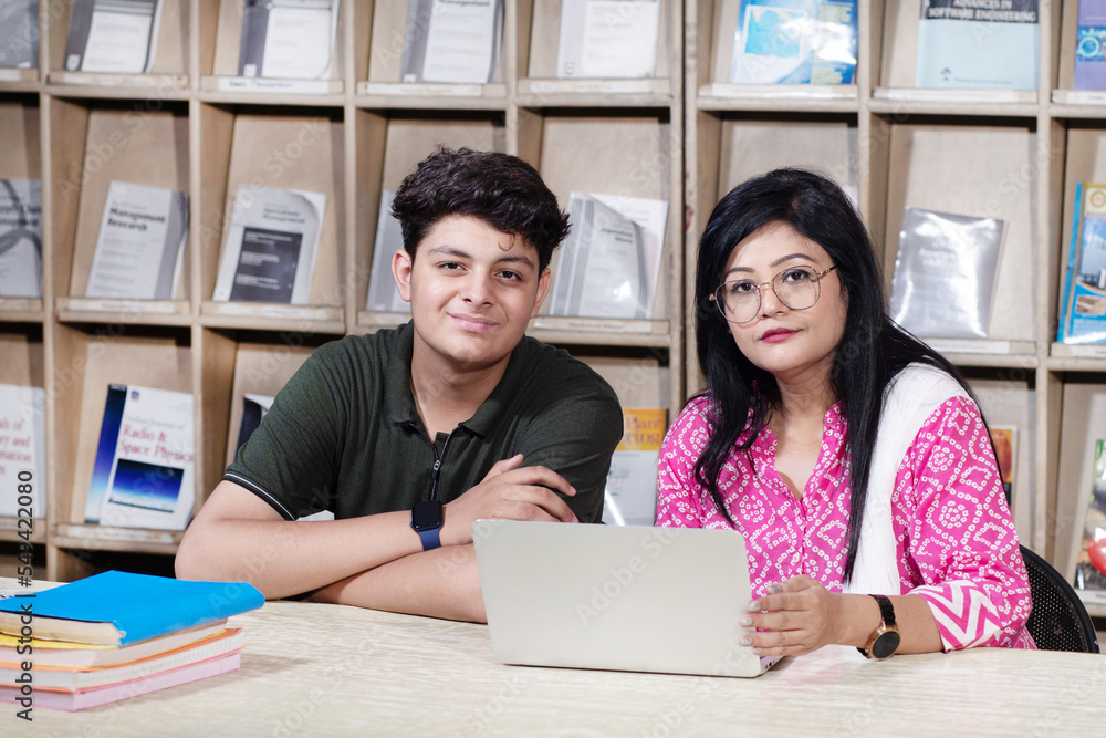 Obraz premium Young indian student boy and female teacher using laptop studying in college library with bookshelf behind. working on assignment or project.