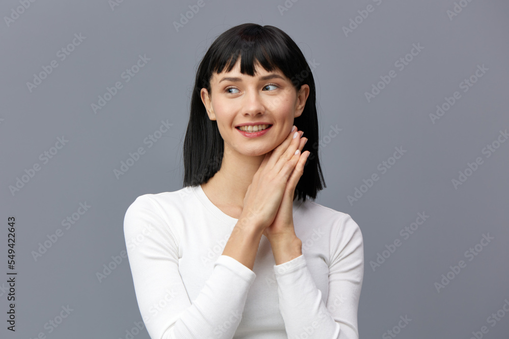 a relaxed woman stands full-face on a dark background in a white tight T-shirt, holds her hands near her face and smiles looking away a relaxed woman stands full-face on a dark background in a white tight T-shirt, holds her hands near her face and smiles looking away