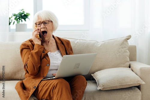 a sad, elderly businesswoman is sitting on a sofa in a cozy apartment in a brown suit working from home and talking on the phone loudly shouts with her mouth wide open