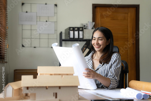 Architecture, building, construction and real estate concept - Happy smiling female architect with blueprint, ruler and living house model on table working at home office. Architect woman.