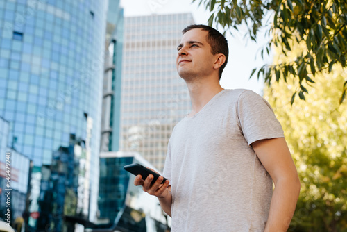 Smiling handsome adult guy holding smartphone standing on city street and looking away