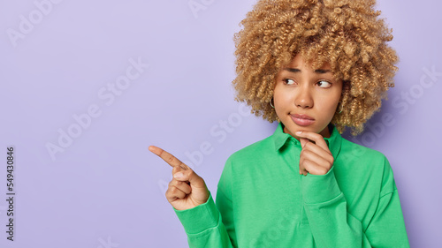 Horizontal shot of serious curly haired woman being deep in thoughts keeps hand on chin indicates index finger aside shows something on left side wears casual green jumper isolated over purple wall