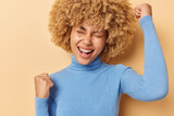 Lucky cheerful young woman with curly bushy hair clenches fists celebrates personal achievements exclaims gladfully feels like winner wears casual blue turtleneck isolated over beige background