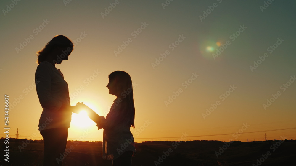 Mom and daughter hold hands, look at each other. Silhouettes at sunset