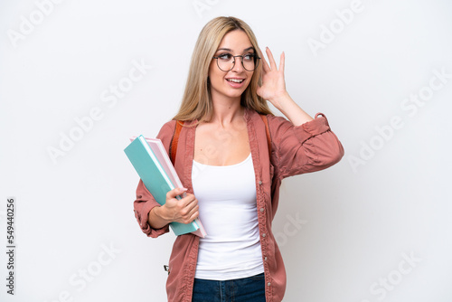 Pretty student blonde woman isolated on white background listening to something by putting hand on the ear