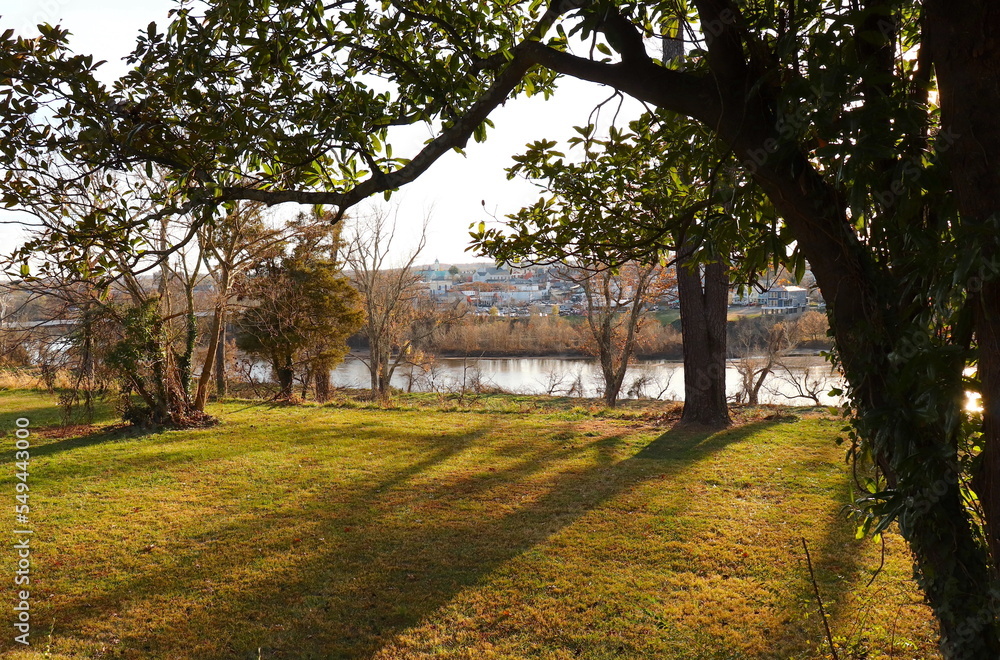 Obraz premium Town Visible Across the River, Pre-Sunset Light Casts Shadows on Grass