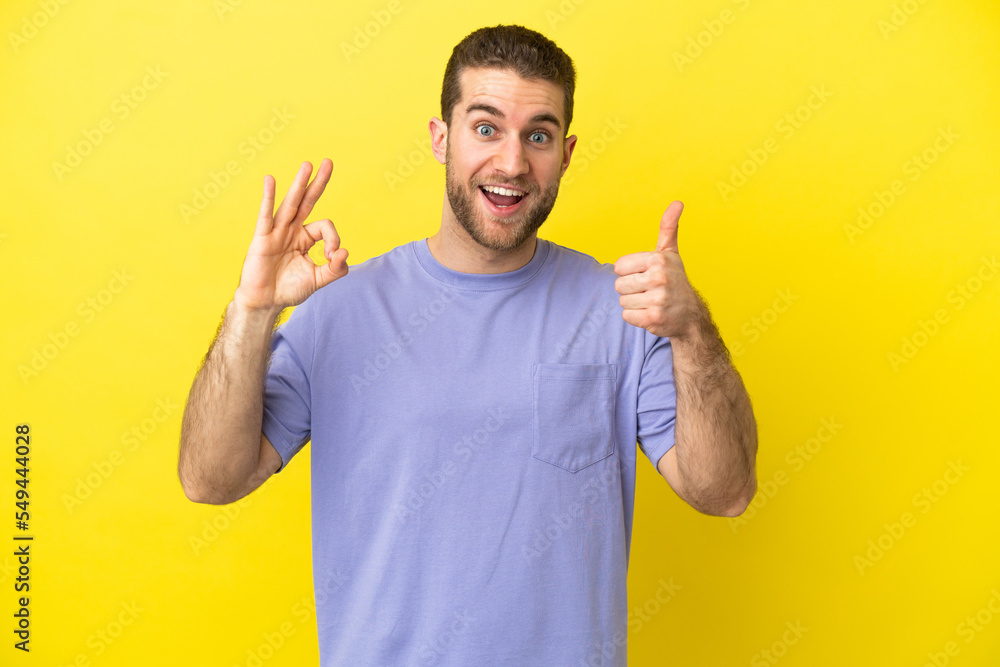 Handsome blonde man over isolated yellow background showing ok sign and thumb up gesture Handsome blonde man over isolated yellow background showing ok sign and thumb up gesture