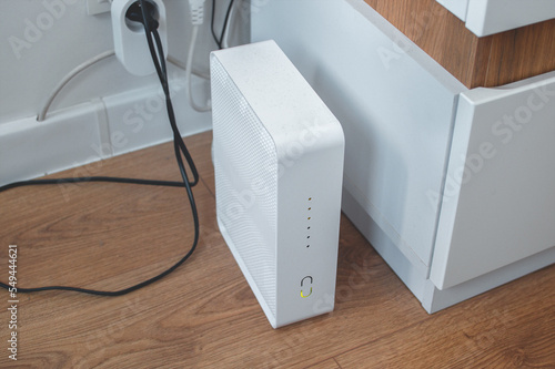 A modern Wi-Fi router plugged into an outlet, white and without antennas, stands on the floor in a home room.