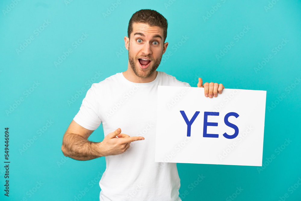 Handsome blonde man over isolated blue background holding a placard with text YES and pointing it Handsome blonde man over isolated blue background holding a placard with text YES and pointing it