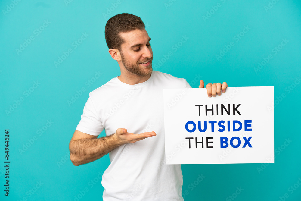 Handsome blonde man over isolated blue background holding a placard with text Think Outside The Box and pointing it Handsome blonde man over isolated blue background holding a placard with text Think Outside The Box and pointing it