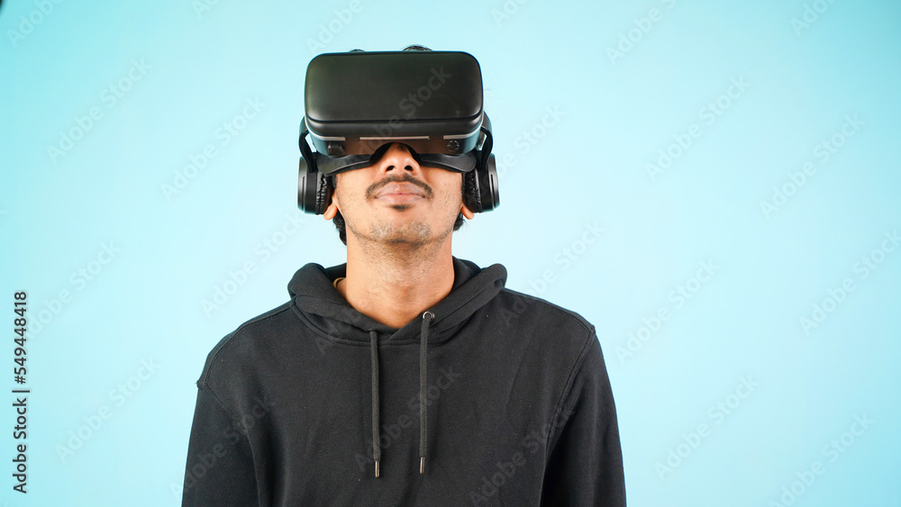 Young Indian Asian man using VR headset, glasses or goggles, isolated over blue colour indoor studio background, future, gadgets, technology, education online, studying, video game concept Young Indian Asian man using VR headset, glasses or goggles, isolated over blue colour indoor studio background, future, gadgets, technology, education online, studying, video game concept