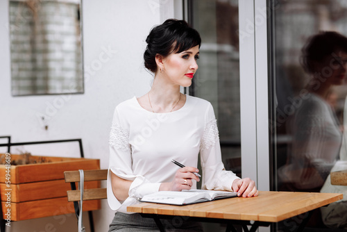 Serious focused smart businesswoman is writing notes on notepad, making agenda on personal organizer. Female in business suit sitting at table at cafe on terrace, work and event planning. photo