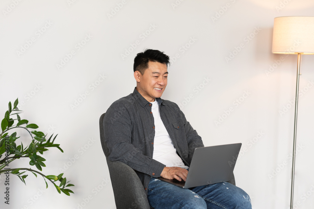 Happy mature japanese man typing on computer, watch video, sits in armchair in living room interior Happy mature japanese man typing on computer, watch video, sits in armchair in living room interior