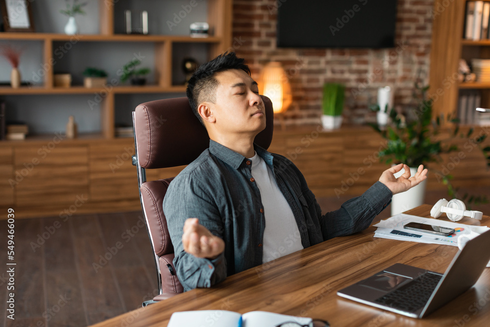Obraz premium Relaxed adult asian man with closed eyes meditation, rests at table with laptop, enjoys silence and comfort