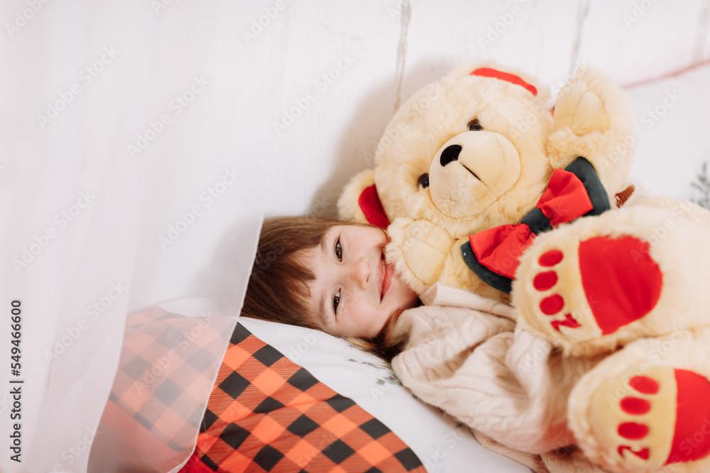 Adorable smiling little girl is lying and playing with teddy bear on bed in room decorated garlands for New Year. Winter weekends. Cozy scene. Christmas holiday atmosphere.