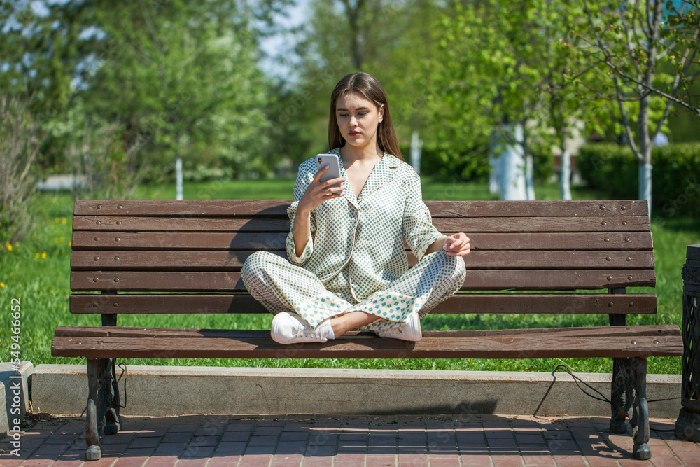 young brunette woman with a phone sits on a bench in a summer park
