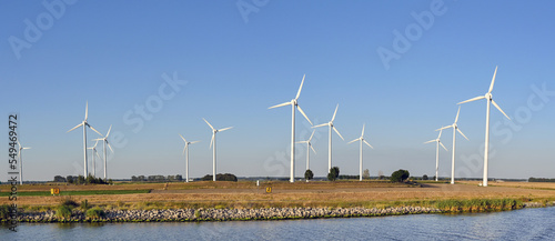 Panoramic view of wind turbines on farmland alongside a river. No people.
