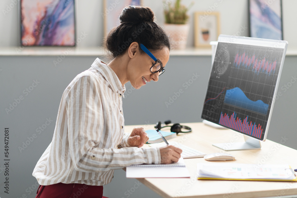 Happy businesswoman taking notes and analyzing indexes, financial charts trading online, graph on pc screen